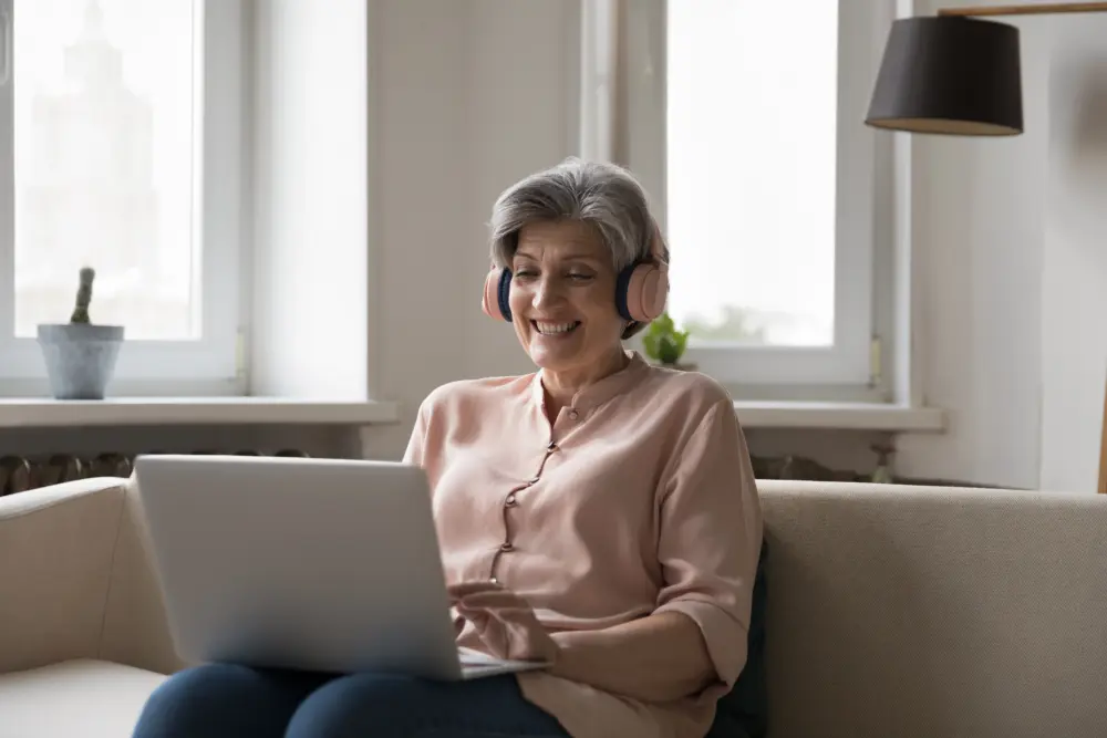 Smiling older woman wearing headphones, taking part in an online counselling session on her laptop at home.